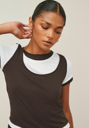 Junge Frau mit glattem Haar und goldenen Creolen, die ein schwarzes ärmelloses Top über einem weißen kurzärmligen Shirt trägt und nach unten schaut, dabei die Hand nahe am Ohr hält.