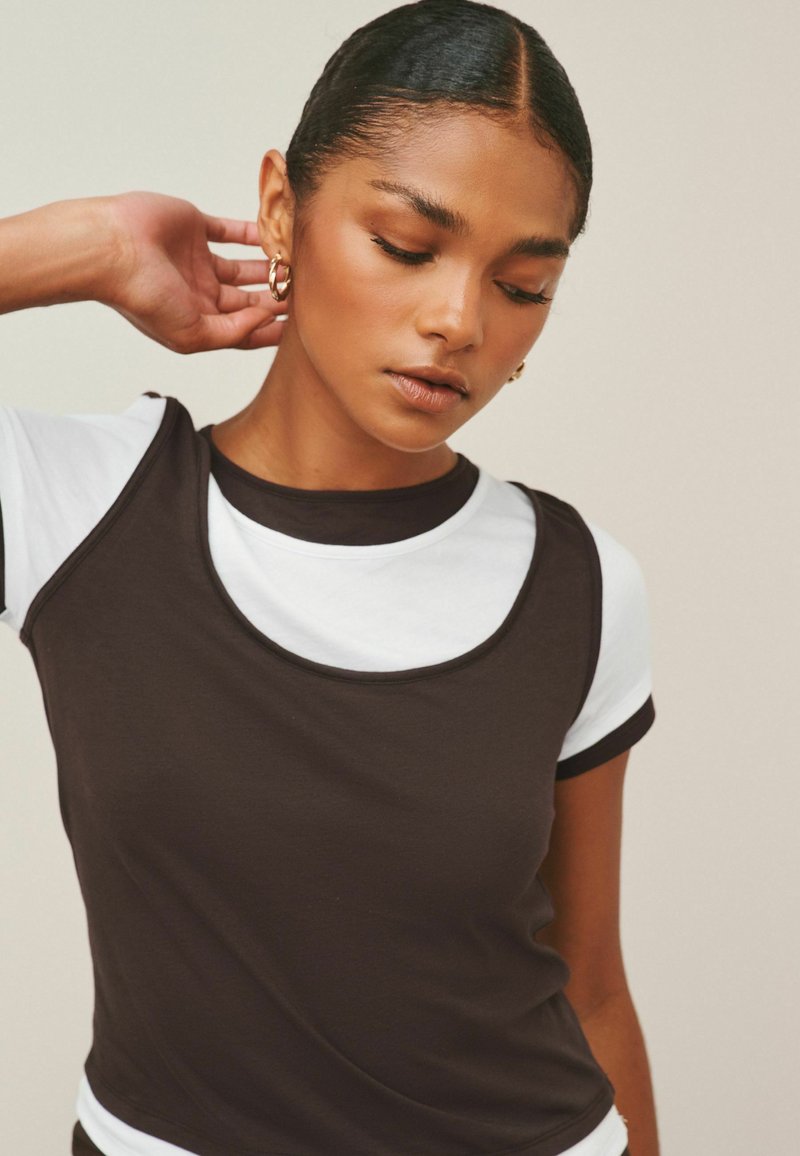 Ung kvinde med glat hår og guldringøreringe, iført en sort ærmeløs top over en hvid kortærmet skjorte, der kigger ned med hånden nær øret.