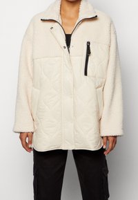 Krämfärgad fleece- och vadderad jacka med en dragkedjeficka, tryckknappar och hög krage. Har vida ärmar och kontrasterande texturer.