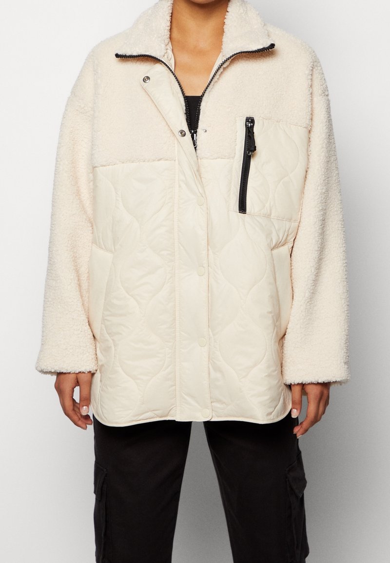 Krämfärgad fleece- och vadderad jacka med en dragkedjeficka, tryckknappar och hög krage. Har vida ärmar och kontrasterande texturer.