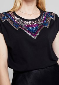 Frau in einem schwarzen Shirt mit einem mehrfarbigen, glitzernden geometrischen Muster am Ausschnitt und einer dünnen goldenen Halskette.