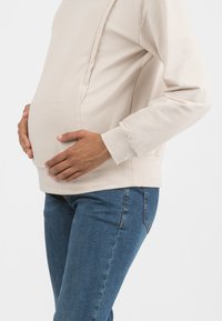 Felpa beige con orlo arrotondato, texture morbida e cuciture laterali, abbinata a jeans skinny blu, con le mani appoggiate su un pancione visibile.