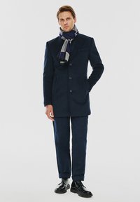 Granatowy wełniany płaszcz o dopasowanym kroju, z dwiema przednimi kieszeniami, zapięcie na guziki, zestawiony z pasującymi spodniami i wzorzystym szalem. Czarne buty eleganckie.