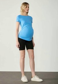 Zila grūtniecības blūze ar izliekta šuvuma dizainu, saskaņota ar melniem šortiem. Modelis valkā baltas sporta kurpes un stāv uz neitrāla fona.