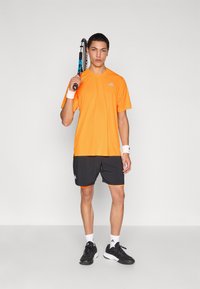 Πορτοκαλί αθλητική μπλούζα, μαύρο σορτς με πορτοκαλί λεπτομέρειες, βραχιόλια καρπού και μαύρα αθλητικά παπούτσια. Το μοντέλο κρατάει ρακέτα τένις.