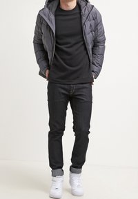 Man som bär en grå puffjacka över en svart skjorta, mörka jeans med uppvikta fållar, och vita högstövlar, står med händerna i fickorna.