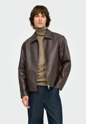 Jeune homme portant une veste en cuir marron, un pull à col roulé beige et un jean bleu, debout devant un fond clair uni.