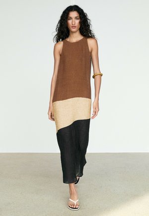 Donna con capelli ricci che indossa un vestito smanicato a blocchi di colore marrone, beige e nero, che cammina a piedi nudi su un pavimento chiaro contro uno sfondo semplice.
