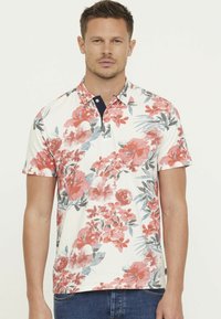 Polo à manches courtes présentant une base blanche avec de grands motifs floraux corail et gris, un col boutonné avec une touche de marine, et une texture douce.