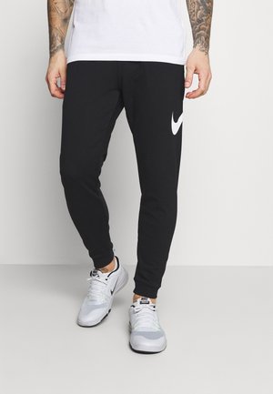 Pantalon de jogging noir avec un logo Nike blanc, présentant une coupe fuselée et des poignets côtelés. Associé à des chaussures de sport blanches.