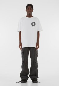 Biely voľný tričko s okrúhlym čiernym grafickým motívom na hrudi, skombinované s voľnými čiernymi cargo nohavicami s viacerými vreckami.