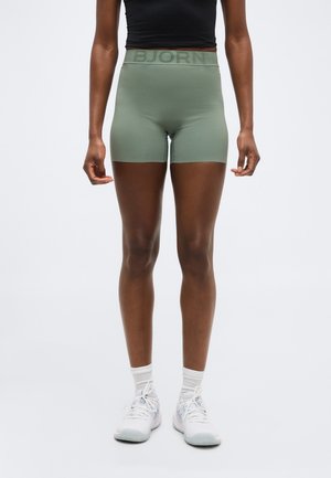 Személy világoszöld, magas derekú rövidnadrágban, melynek derékrészén a „BJORN” felirat olvasható, fekete rövid felsőben, fehér zokniban és fehér sportcipőben, egyszínű háttér előtt.