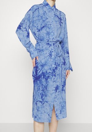 Femme portant une robe chemise bleue tie-dye à manches longues et boutonnée, avec une ceinture à la taille, debout avec une main sur la hanche.