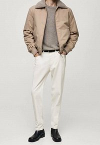 Giacca bomber marrone con colletto in pelliccia, maglione grigio, pantaloni bianchi e stivali neri alla caviglia. Materiale liscio, toni neutri, vestibilità casual.