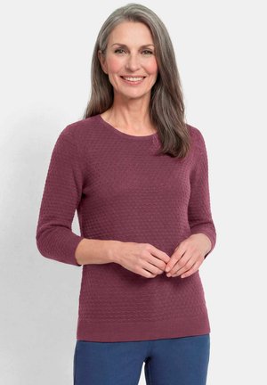 Glunderende vrouw van middelbare leeftijd met lang grijs haar, die een gestructureerde mauve trui met lange mouwen en een blauwe broek draagt, staand tegen een effen achtergrond.
