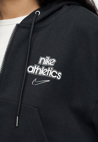 Μαύρο φούτερ Nike Athletics με φερμουάρ, κορδόνια και λευκό λογότυπο στο στήθος, φορεμένο πάνω από λευκό ριμπ μπλουζάκι.