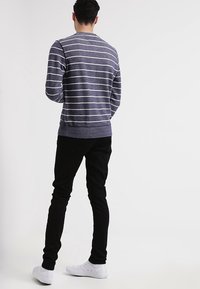 Man in een marineblauwe en witte gestreepte lange mouwen shirt, zwarte broek en witte sneakers, staande met zijn gezicht weg van een witte achtergrond.