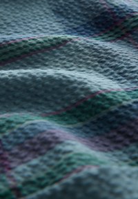 Tekstureret stof i nuancer af blå, grøn og lilla med et ternet mønster og en blød, bølgende overflade.