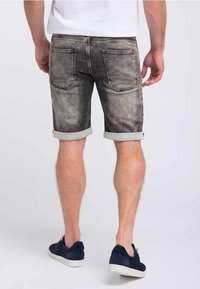 Pantalones cortos de mezclilla gris con textura desgastada, dobladillos enrollados y dos bolsillos traseros, combinados con zapatillas oscuras en un fondo neutro.