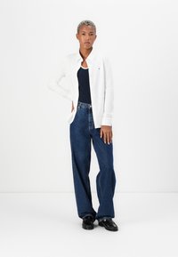 Witte button-up shirt over een marineblauwe tanktop, gecombineerd met losse blauwe jeans en zwarte schoenen. Schoon ontwerp met minimale accenten.