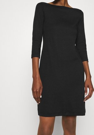 Femme portant une robe noire mi-longue aux genoux avec des manches trois-quarts et un col bateau, se tenant devant un fond blanc uni.