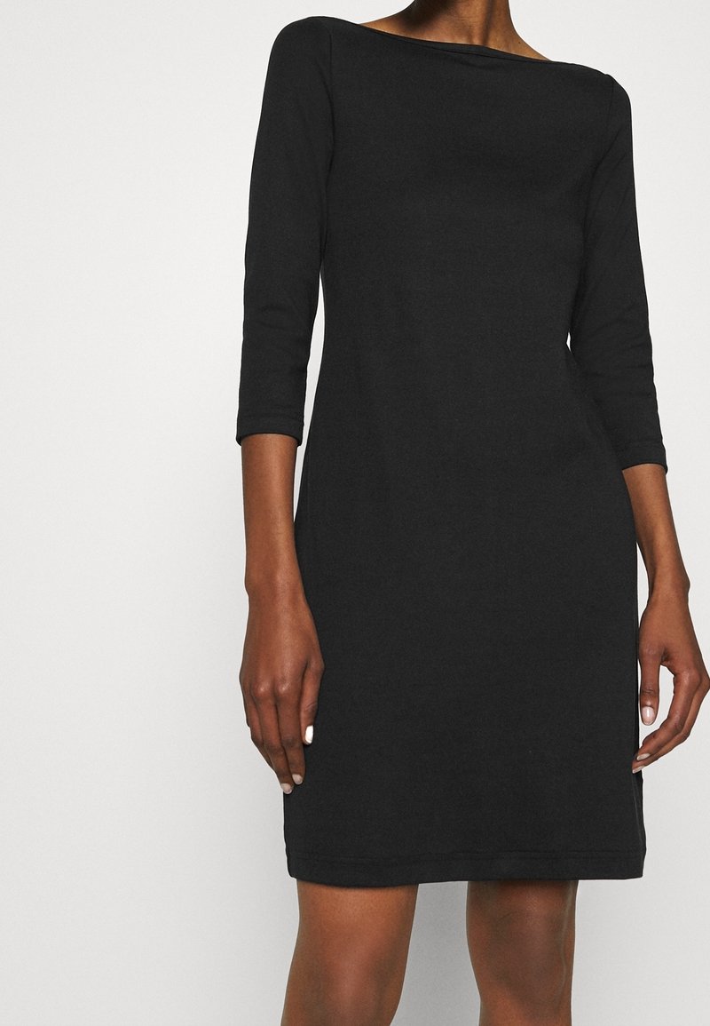 Femme portant une robe noire mi-longue aux genoux avec des manches trois-quarts et un col bateau, se tenant devant un fond blanc uni.