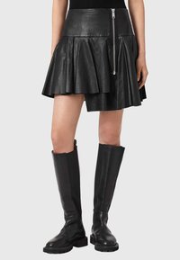 Γυναίκα που φοράει μαύρη ασύμμετρη πλισέ δερμάτινη φούστα με ασημένιο φερμουάρ και μαύρες δερμάτινες μπότες μέχρι το γόνατο.