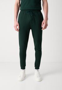 Mørkegrønne joggebukser laget av mykt stoff, med elastisk liv med snøring og tettsittende mansjetter ved anklene.