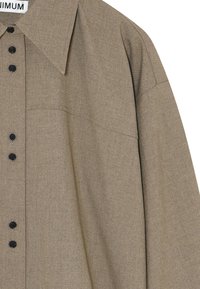 Camisa de botones de color marrón claro con un gran cuello, botones negros y una tela texturizada. Presenta un corte holgado y detalles de costura mínimos.