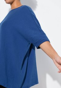 Top bluaggio largo realizzato in tessuto testurizzato, con scollo ampio, maniche a tre quarti e spacchi laterali per facilitare i movimenti.