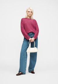 Νεαρή γυναίκα με ματζέντα πουλόβερ, φαρδιά μπλε τζιν, κρατώντας μια μικρή λευκή τσάντα, στέκεται μπροστά σε ένα απλό ανοιχτό φόντο.