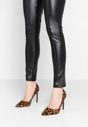 Benen met zwarte leren broek en hoge hakken met luipaardprint en puntige neus op een witte vloer met witte muur op de achtergrond.