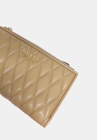 Portafoglio beige trapuntato con logo dorato "GUESS EST. 1981". Texture in pelle liscia, chiusura con zip, motivo a cuciture diagonali e bordi arrotondati.