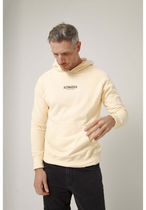 Hombre con pelo gris corto que lleva una sudadera con capucha color crema con el texto "ALTONADOCK it's your way", mirando hacia abajo con una mano levantada.