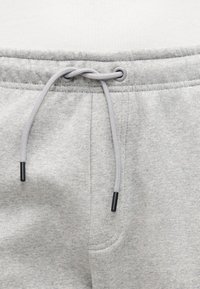 Ceinture de pantalon de survêtement gris clair avec cordon gris noué, embouts en plastique noirs, et couture détaillée autour de la braguette et des poches.