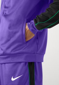 Fialová športová bunda s čiernymi rukávmi, s zelenými akcentmi. Obsahuje bočnú kapsu, vyrobená z hladkej látky. Viditeľné logo Nike.