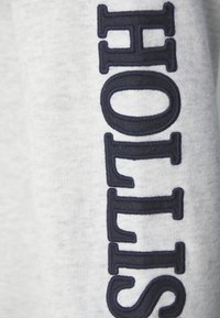 Tela de sudadera gris con texto bordado en azul marino oscuro que deletrea "HOLLIS" verticalmente a lo largo de la manga, con una textura suave.