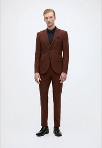 Bruin op maat gemaakt pak met een slim fit, gecombineerd met een zwart overhemd en een bijpassend pochet. Het pak heeft twee knopen en notch revers.
