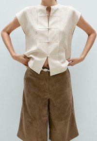 Lniana bluzka z krótkim rękawem w jasnobeżowym kolorze, z kołnierzem typu mandarynka i zapinaną na guziki listwą, w parze z brązowymi spodniami culottes zamszowymi o gładkiej fakturze.