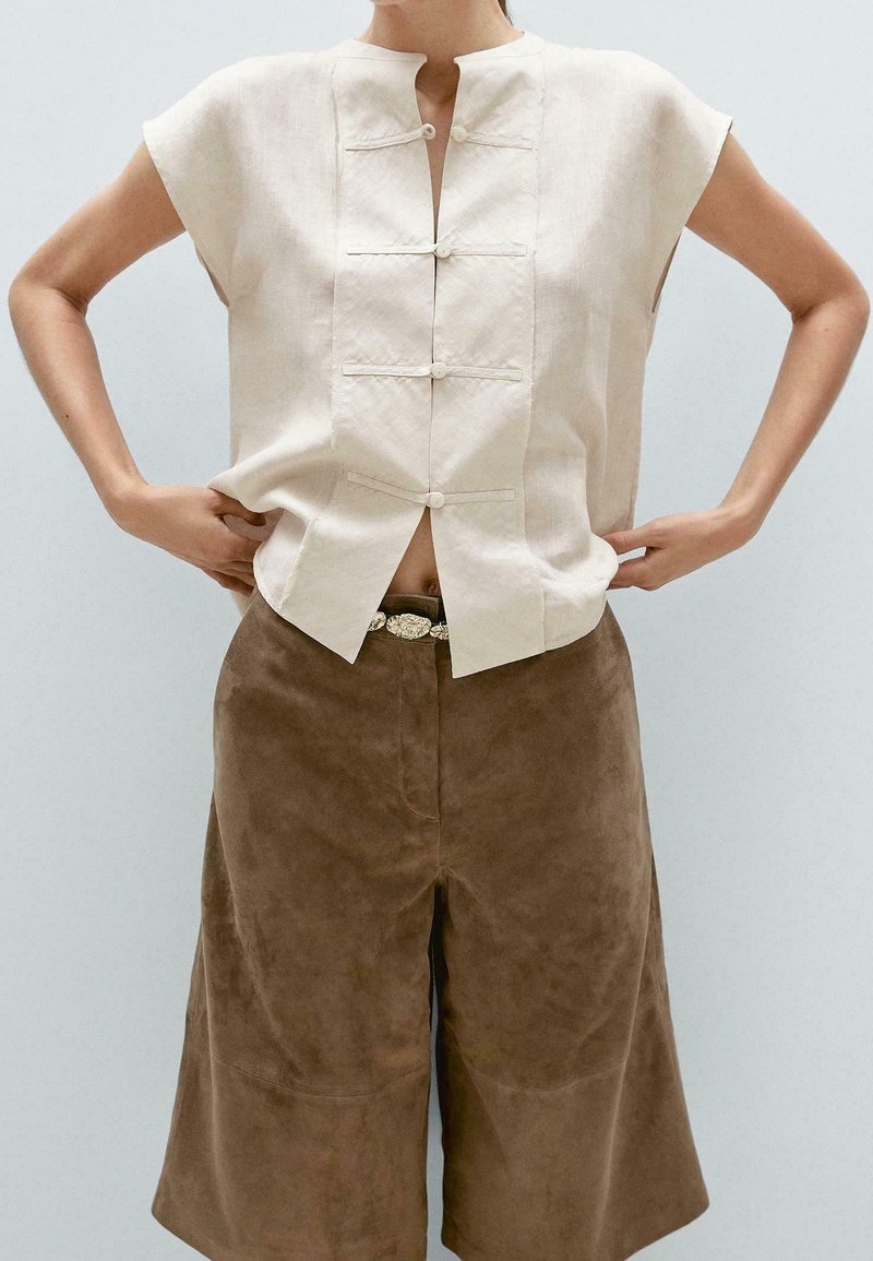 Lniana bluzka z krótkim rękawem w jasnobeżowym kolorze, z kołnierzem typu mandarynka i zapinaną na guziki listwą, w parze z brązowymi spodniami culottes zamszowymi o gładkiej fakturze.
