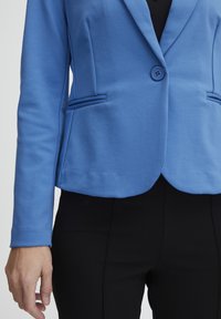 Modrý blazer se přiléhavým střihem, zapínáním na jeden knoflík, dvěma předními kapsami a texturovanou látkou, v kombinaci s černými kalhotami.