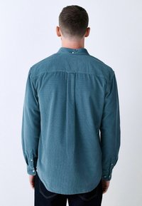 Cămașă cu butoni, din terciote teal, cu pliseu la spate, guler și manșete cu butoni. Țesătură texturată cu un model subtil de nervuri.
