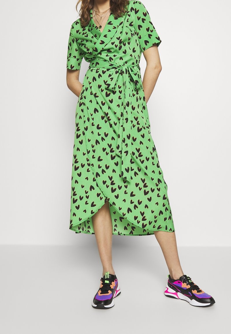 Femme portant une robe portefeuille verte avec des motifs de cœurs noirs et des baskets colorées noires, violettes, roses et orange, debout devant un fond uni.