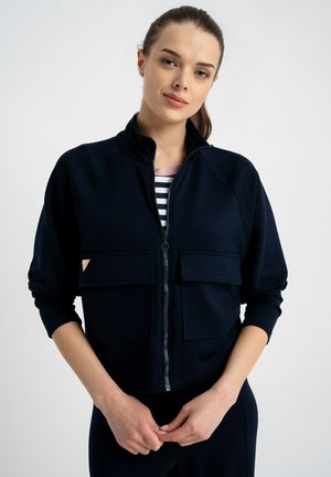 Kobieta z związanymi włosami, ubrana w czarną kurtkę na suwak na paskowanej koszulce, trzymająca ręce splecione przed sobą na jednolitym tle.