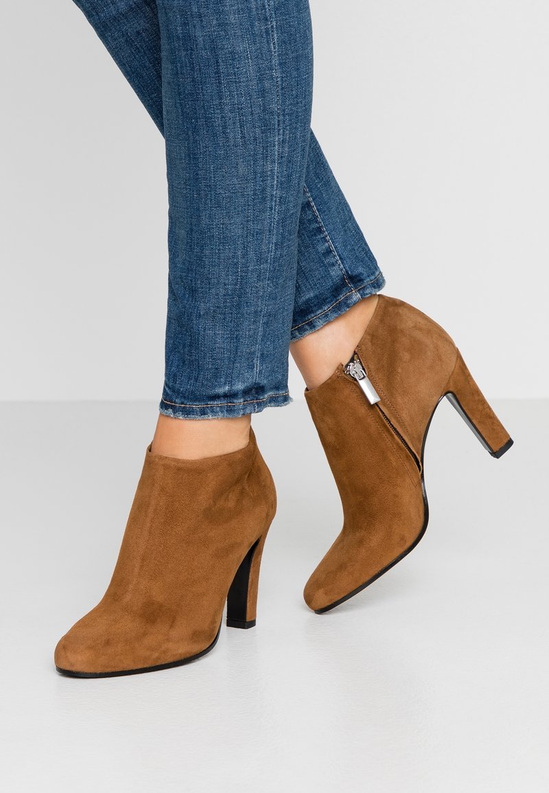 Brune rågummistøvler med spidse tæer og høje hæle; har en lynlås i siden og en glat, jævn tekstur. Bruges med mørkeblå jeans.