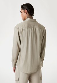 Camisa de manga larga, de color beige claro, con una textura suave, collar tipo button-down y mangas con puños. Presenta un dobladillo curvado y un pliegue en la parte posterior.