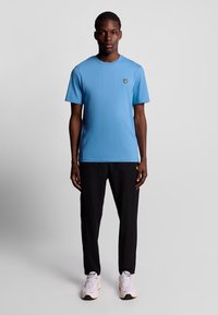 Ljusblå bomulls t-shirt med korta ärmar, rund halsringning och en liten gul fjärilslogotyp på bröstet. Svarta byxor och ljusa sneakers.