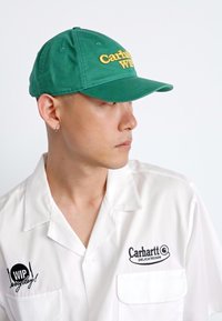 Casquette en coton verte avec "Carhartt WIP" brodé en jaune, dotée d'une visière courbée. Le modèle porte une chemise blanche avec des accents noirs.