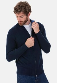 Pull à fermeture éclair bleu marine en tissu tricoté, avec un col montant, des poignets côtelés et une coupe ajustée, porté sur une chemise blanche.