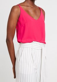 Haut rose vif sans manches avec un col en V, en tissu lisse. Associé à un pantalon blanc à fines rayures verticales et une taille nouée.